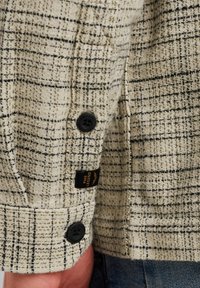 Veste à carreaux beige en tissu texturé, ornée de boutons noirs, d'une étiquette sur la manche et d'un poignet à motif tartan.