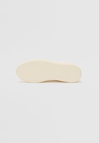 Sole of sneaker in off-white with a slightly textured surface and zigzag pattern for grip. Rounded shape with a smooth edge.