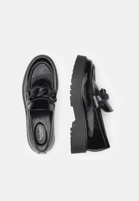 Mocassins en cuir verni noir avec une semelle épaisse et un accent décoratif en chaîne. Texture lisse avec un bout arrondi et un petit talon.