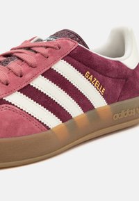 Tmavě růžová semišová teniska s bílými koženými pásky, zlatým logem „GAZELLE“ a podrážkou z gumy v přírodní barvě. Texturované tkaničky a polstrovaný kotník.