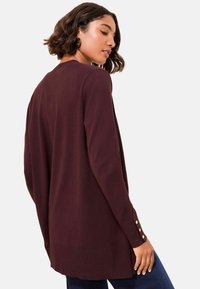Long cardigan bordeaux avec une finition texturée, présentant une coupe droite, un col rond et trois accents de boutons dorés sur chaque manche.