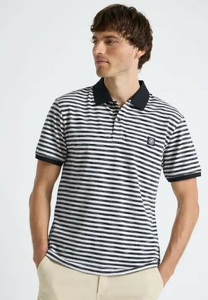 Homme portant un polo rayé noir et blanc avec col noir et pantalon beige, debout les mains dans les poches, regardant vers la gauche.