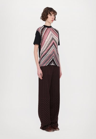 Top de punto con patrón de zigzag en rosa y negro, combinado con pantalones amplios marrones con un diseño de zigzag. La modelo está de pie sobre un fondo neutro.