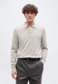 Νεαρός άνδρας με σγουρά μαλλιά, ντυμένος με μπεζ μακρυμάνικο πόλο πουκάμισο και σκούρο παντελόνι, στέκεται με το ένα χέρι στην τσέπη.