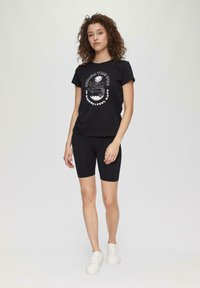 Czarna bawełniana koszulka z grafikiem z przodu, zestawiona z czarnymi szortami rowerowymi. Model nosi białe sneakersy. Gładkie tło.