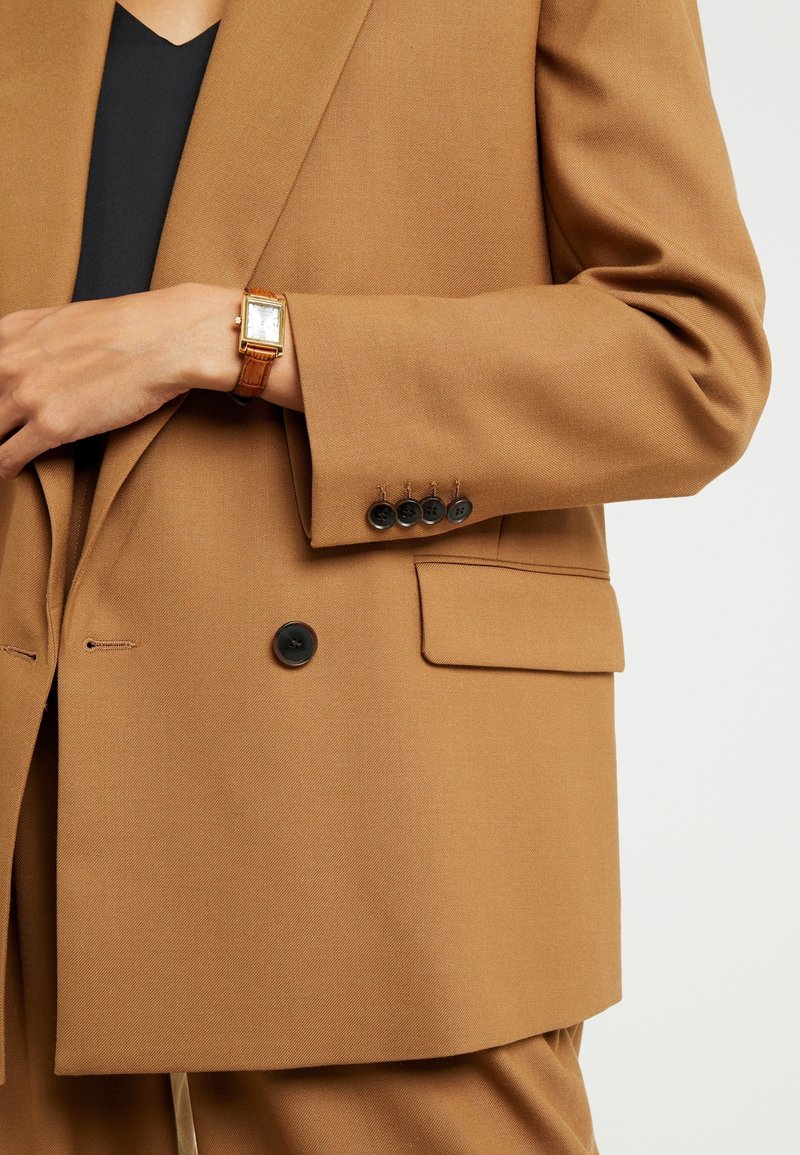 Personne portant un blazer marron et un haut noir, avec une montre-bracelet rectangulaire dorée au poignet gauche, gros plan sur le torse et le bras.