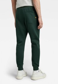 Pantalones deportivos verdes, hechos de una tela suave, con un bolsillo trasero, puños elásticos y un pequeño detalle de logo. Llevados con zapatillas blancas.