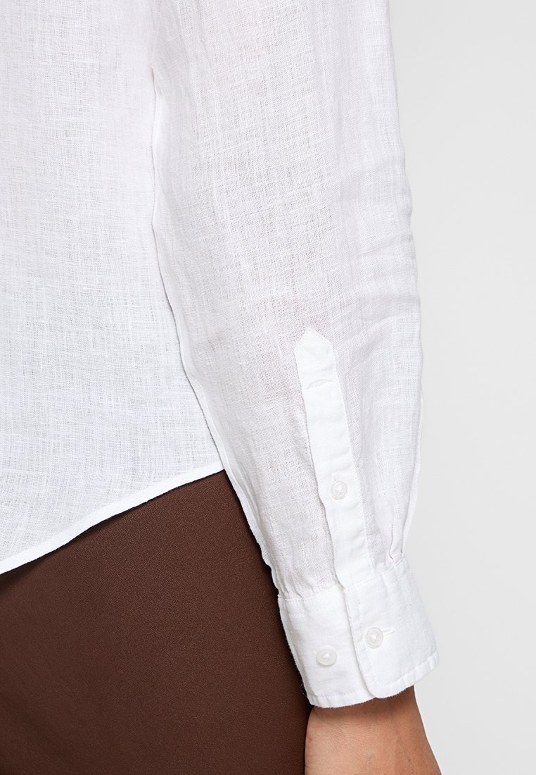 Biała lniana koszula zbliżenie, z teksturowanym materiałem, zapięciem na guziki przy mankietach i luźnym krojem. Mankiet ma dwa guziki i spiczasty kształt.