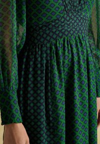 Zielona i granatowa sukienka z geometrycznym kwiatowym wzorem, przezroczystymi długimi rękawami, dopasowaną talią i marszczonym dołem. Teksturowany materiał z detalami warstwowymi.