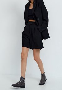 Blazer nero con un bottone singolo, abbinato a un top corto nero e pantaloncini. Stivaletti neri con materiale lucido e lacci completano il look.