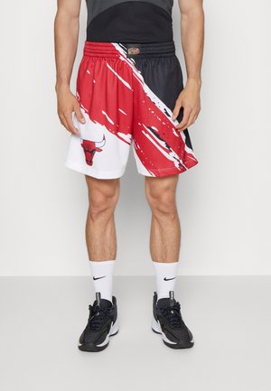 Homme portant un short de basket Chicago Bulls noir et rouge avec des chaussettes blanches Nike et des baskets noires, debout devant un fond uni.