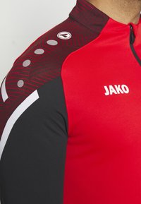 Rode en zwarte sporttop met hoge kraag, met een getextureerd grafisch patroon, witte accenten en ronde grijze details op de schouders.