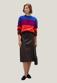 Bunter gestreifter Pullover in Blau, Lila, Rot und Braun; kombiniert mit einem dunkelbraunen Leder-Midirock; schwarze Schuhe; grüne Tasche.