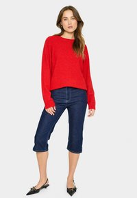 Roter Strickpullover mit Rundhalsausschnitt, kombiniert mit dunkelblauen, hüftlangen Jeans und schwarzen, spitz zulaufenden Ballerinas, die dezente Details aufweisen.
