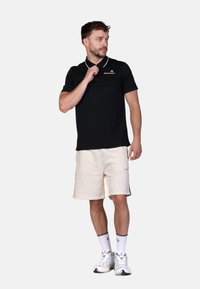 Polo shirt nera con accenti bianchi e logo, abbinata a pantaloni corti color crema. Il modello indossa calzini bianchi e sneakers; look sportivo e casual.