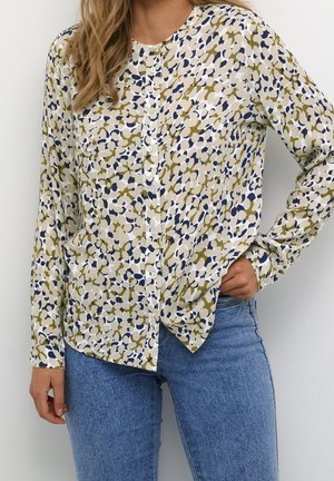 Kobieta ubrana w bluzkę na guziki z długim rękawem o abstrakcyjnym wzorze w kolorach beżowym, granatowym i oliwkowym, zestawioną z jasnoniebieskimi dżinsami.