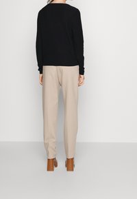 Eine Person trägt einen schwarzen Pullover, beigen Hosen und braunen Absatzschuhen und steht mit dem Rücken zu einem schlichten weißen Hintergrund.