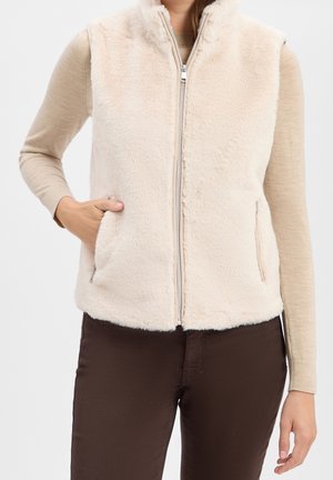 Beige, pluizig vest met rits gedragen over een beige top met lange mouwen, gecombineerd met donkerbruine broek, hand nonchalant in het vestzakje.