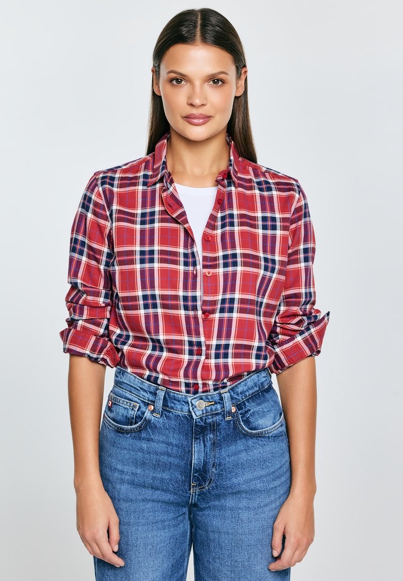 Czerwona flanelowa koszula na guziki w kratę z niebieskimi i białymi akcentami, z podwiniętymi rękawami i klasycznym kołnierzykiem, zestawiona z wysokim stanem niebieskich dżinsów.