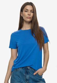 Sieviete ar gariem brūniem matiem, ģērbta vienkāršā zila T-kreklā ar īsām piedurknēm un zilu džinsu, ar vienu roku kabatā uz balta fona.