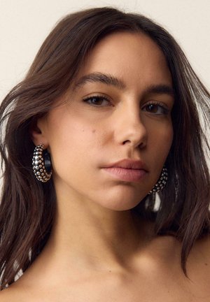 Giovane donna con capelli castani di media lunghezza che indossa grandi orecchini a cerchio neri rotondi con borchie dorate, sfondo neutro.