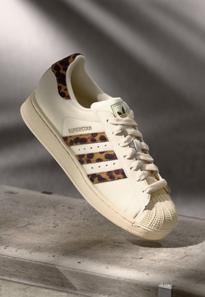Baskets blanches Adidas Superstar avec motif léopard sur les bandes et la languette de talon, bout texturé et semelle couleur crème sur surface grise.