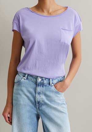 Kobieta w lawendowym t-shircie z krótkim rękawem i kieszonką, włożonym w jasnoniebieskie, wysokie spodnie jeansowe, z jedną ręką w kieszeni.