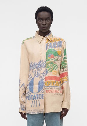 Henkilö, joka käyttää beigeä napillista paitaa, jossa on värikkäitä vintage-tyylisiä maatilan ja perunan kuvioita, yhdistettynä vaaleansinisiin farkkuihin.