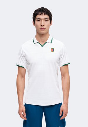 Mann trägt weißes kurzärmeliges Poloshirt mit grünem Rand und Logo auf der Brust, kombiniert mit blauen Shorts, steht vor neutralem Hintergrund.