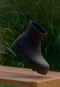 Bota de goma negra de longitud media con punta redondeada, textura mate y suela gruesa, que cuenta con una cremallera en la parte trasera para facilitar su uso.