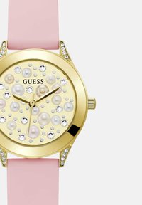 Kuldne ümmargune kell, mille nägu kaunistavad kristallid ja pärlid, roosa rihm ning "GUESS" logo kellakuval 12 kohal.