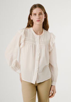 Blusa de un beige claro hecha de una tela suave, con hombros con volantes, un cierre delantero de botones y detalles de pliegues verticales a lo largo del cuerpo.