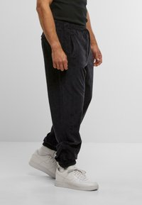 Man wearing textured black sweatpants and white Nike sneakers standing against plain gray background.