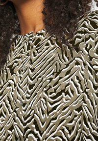 Camisa estampada con un diseño abstracto en verde oliva y blanco, con un cuello abotonado y una tela texturizada.