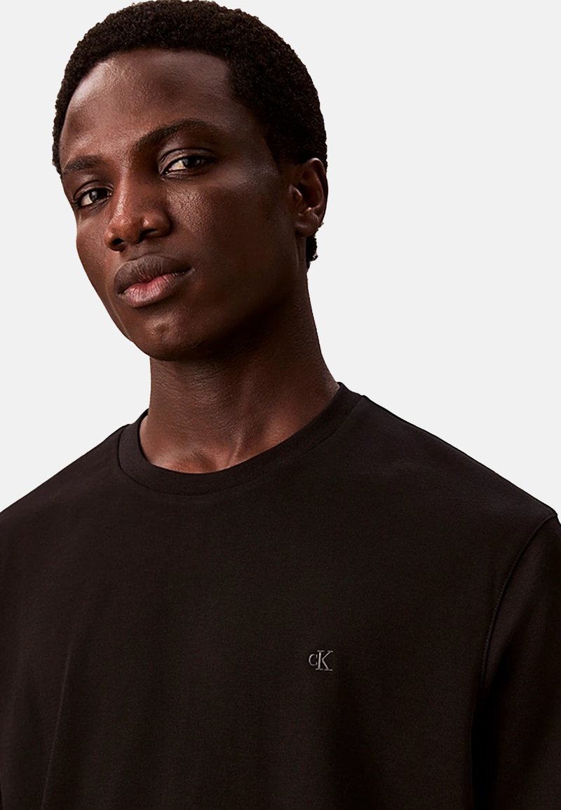 Zwarte crew-neck t-shirt met korte mouwen, voorzien van een klein geborduurd logo aan de bovenlinkerkant. Glad stoffen textuur.