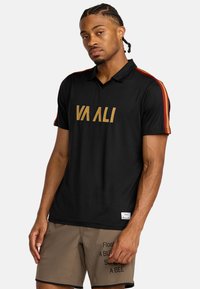Zwarte polo met V-hals en gouden tekst "VA ALI", met rode en oranje strepen op de mouwen. Glad stoffen textuur.