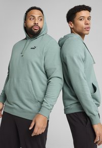 Hoodie vert clair en tissu doux, doté d'une poche kangourou, de poignets côtelés et d'un logo noir sur la poitrine. Deux modèles dos à dos.