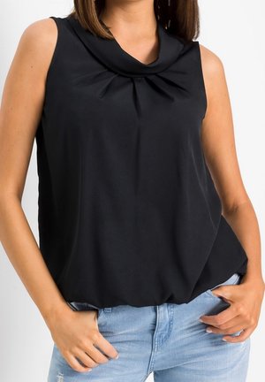 Kobieta w czarnej, bezrękawnikowej bluzce z plisowanym, marszczonym dekoltem oraz jasnoniebieskich dżinsach, z rękami w kieszeniach, na białym tle.