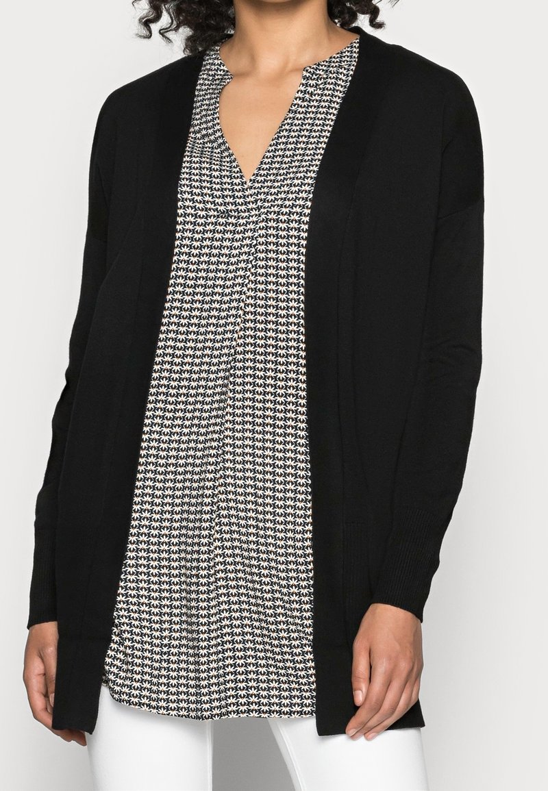 Femme portant un cardigan noir ouvert sur un chemisier à col en V à motif noir et blanc et un pantalon blanc.