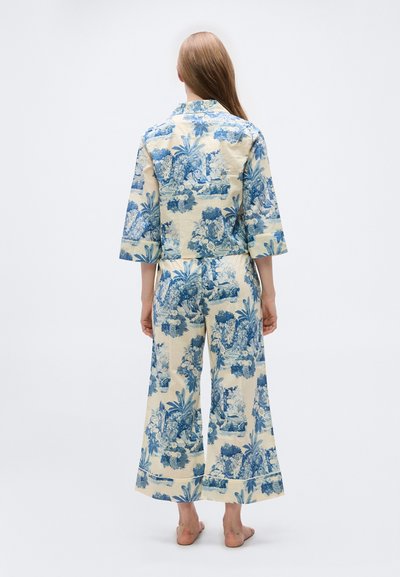 Γυναίκα με μακριά μαλλιά που φοράει εκρού και μπλε φαρδιά παντελόνια με λουλουδάτο μοτίβο και ταιριαστό κοντό σακάκι, όρθια ξυπόλυτη με την πλάτη στραμμένη.