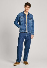 Chaqueta de mezclilla azul con bolsillos frontales, cierre de botones y cinturilla. Combinada con unos vaqueros de mezclilla azul a juego y zapatos de ante marrones.