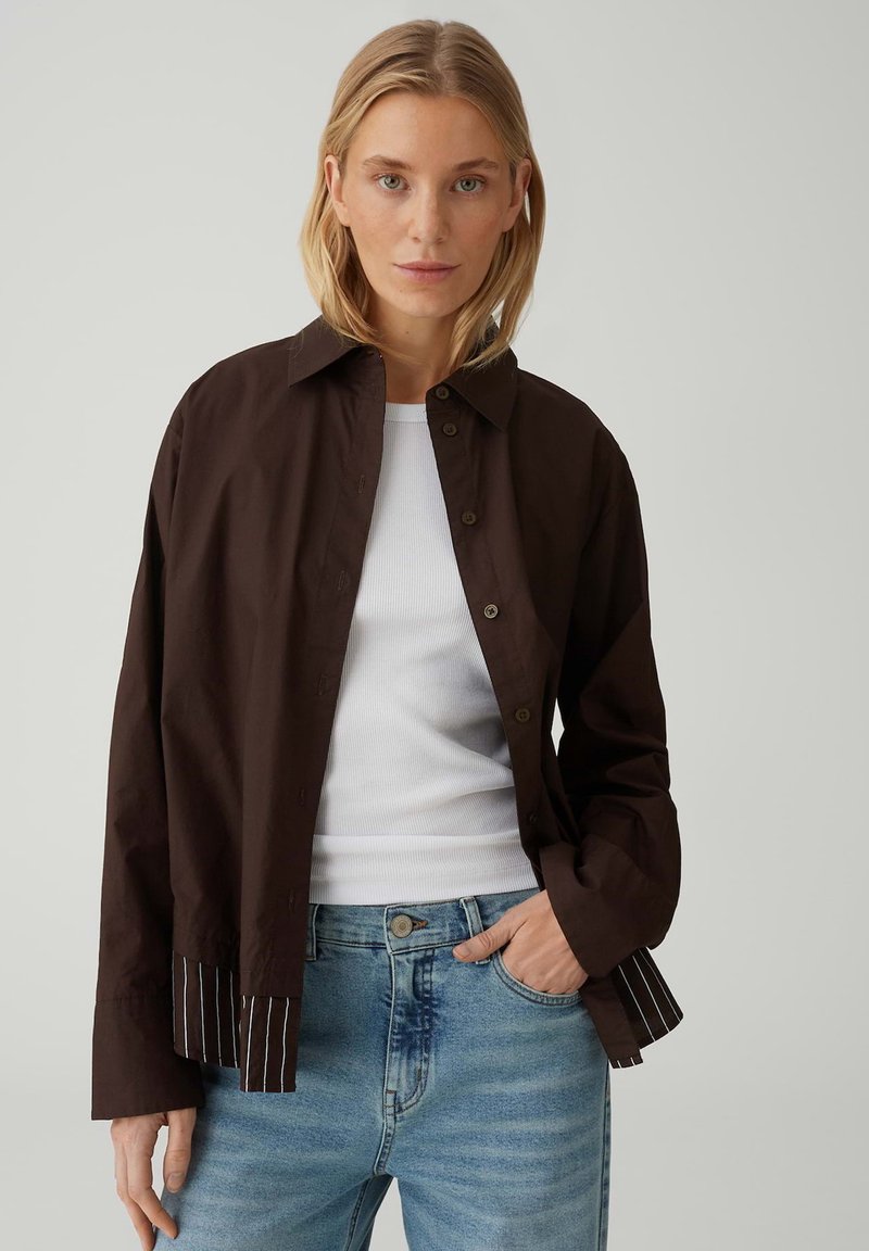 Blonde Frau, die ein offenes dunkelbraunes Hemd über einem weißen, gerippten Tanktop und hellblauen Jeans trägt, vor einem schlichten Hintergrund stehend.