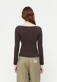 Top maro cu mânecă lungă și guler larg, având un mic logo în formă de triunghi pe spate. Textură netedă și design ajustat.