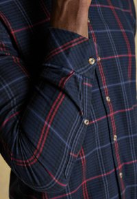Koszula w kratkę w kolorze granatowym z czerwonymi i niebieskimi wzorami, z guzikami z przodu oraz zaokrąglonymi mankietami z widocznymi szwami i guzikami. Teksturowany materiał.