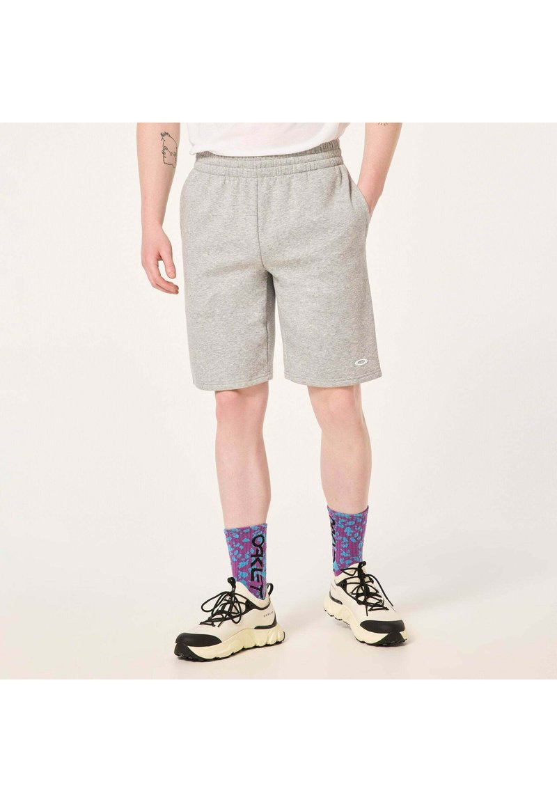 Graue Sportshorts mit elastischem Bund und Seitentaschen, kombiniert mit lila und blauen gemusterten Socken sowie schwarz-weißen Sneakern.