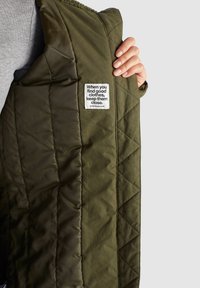 Main tenant une veste matelassée vert olive ouverte, montrant une étiquette blanche avec le texte : « Quand tu trouves de bons vêtements, garde-les près de toi. »