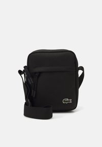 Geantă din material textil negru, cu formă pătrată, închidere cu fermoar, curea ajustabilă și logo brodat verde. Textură netedă, design minimalist.