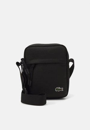 Black fabric bag with a square shape, zip closure, adjustable strap, and green embroidered logo. Smooth texture, minimalist design.
