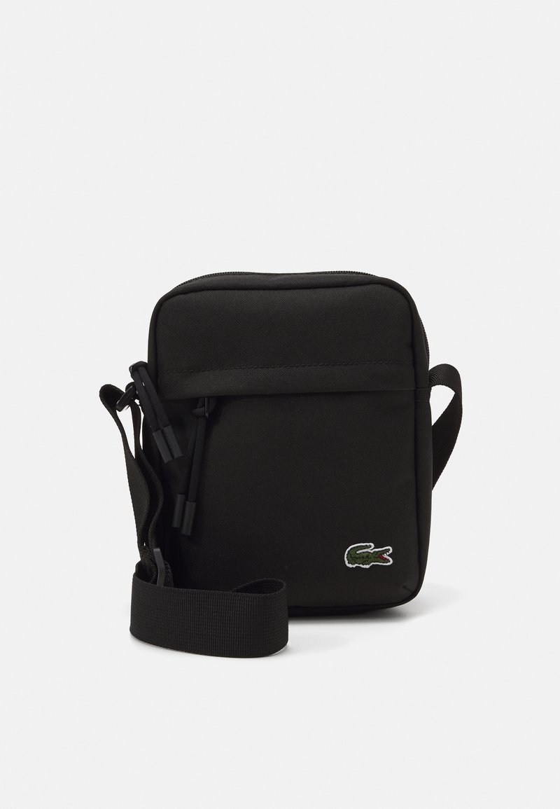 Geantă din material textil negru, cu formă pătrată, închidere cu fermoar, curea ajustabilă și logo brodat verde. Textură netedă, design minimalist.