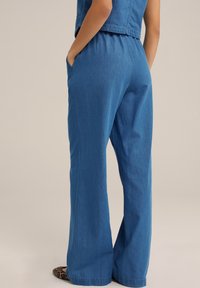 Žena v volných modrých denimových kalhotách a odpovídajícím bezramínkovém topu, s rukama v kapsách, stojící na neutrálním pozadí.